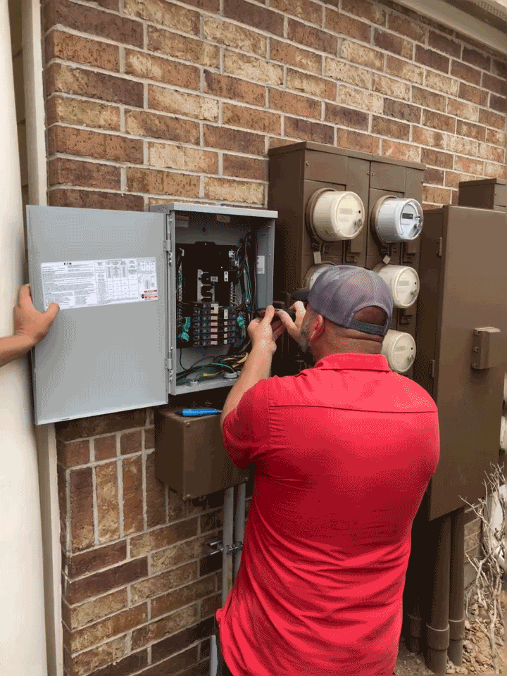 Electrician working on an outdoor electrical panel beside utility meters, performing wiring and circuit maintenance on a brick wall.