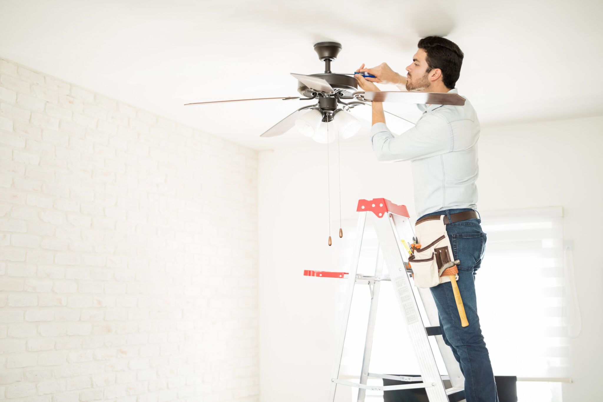 Electrician standing on a ladder while installing a ceiling fan in a bright, modern room.