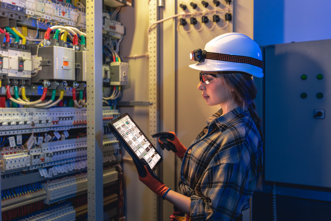 Electrical technician inspecting a control panel with a tablet, wearing a hard hat, safety glasses, and protective gloves.