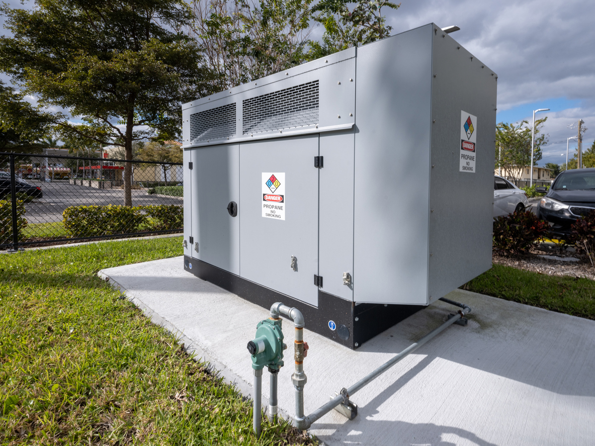 Commercial propane standby generator on a concrete pad with gas piping visible and a parking lot in the background.