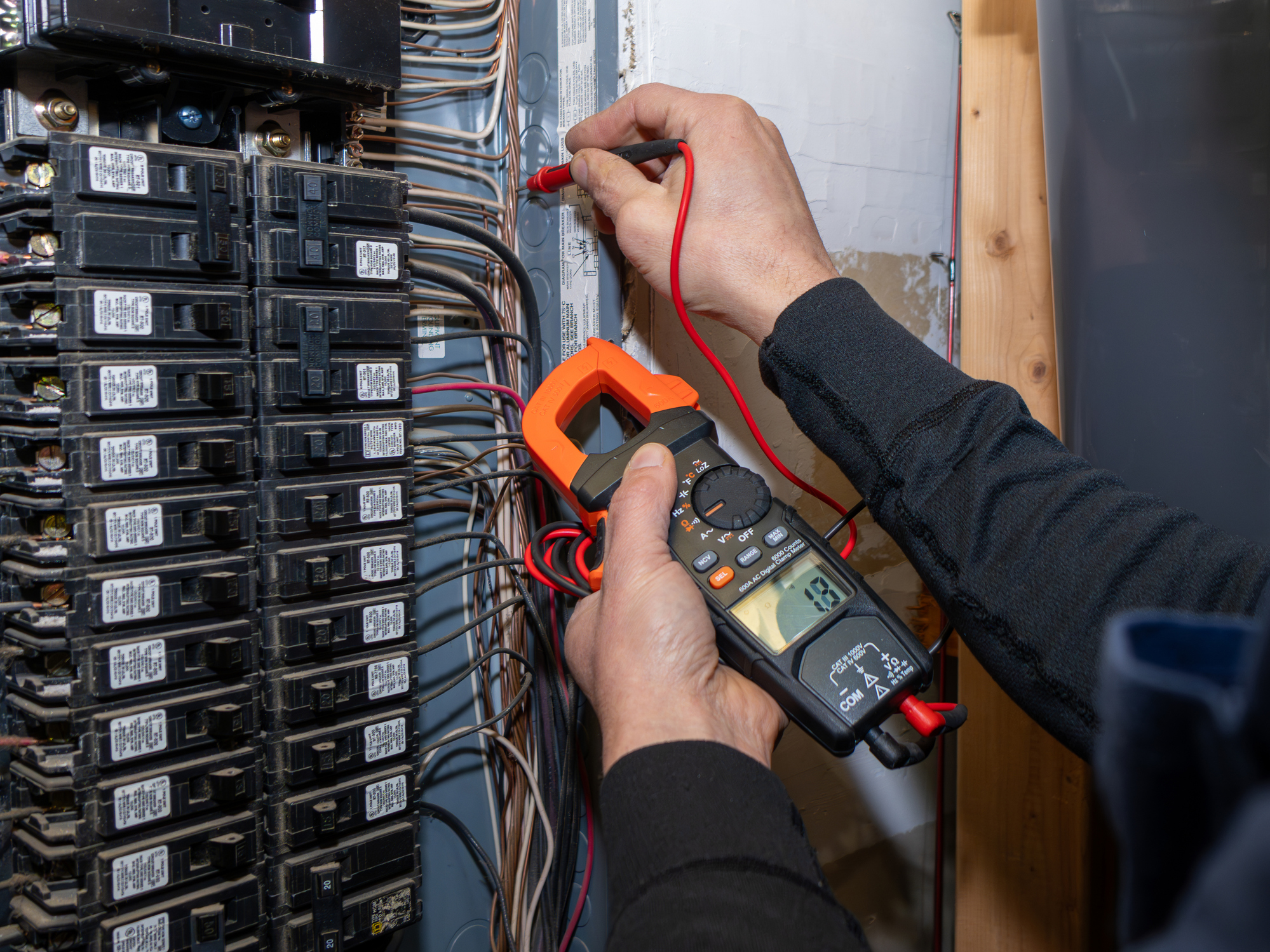 An electrician's hands using an orange digital clamp meter to test voltage inside an open home electrical panel.